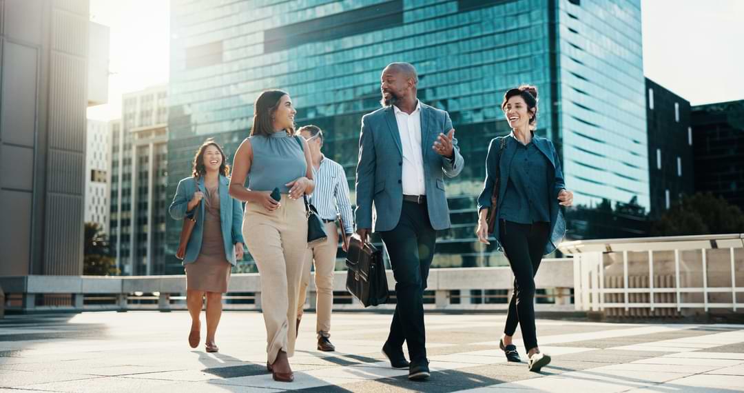 A diverse group of professionals in business attire walks outside a modern office building, smiling and engaged in conversation under bright sunlight.